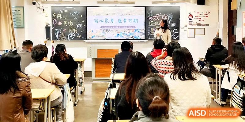 An instructor stands by a chalkboard and screen, presenting a lecture to a classroom full of seated students at Zhengzhou No.19 High School.