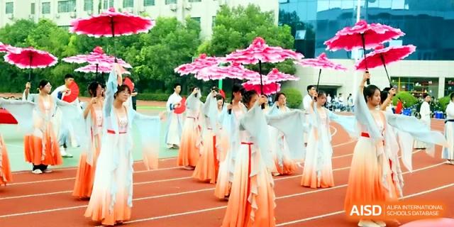 Performers in traditional light-blue and orange flowing dresses carry pink flower props on an outdoor track at Zhengzhou No.19 High School.