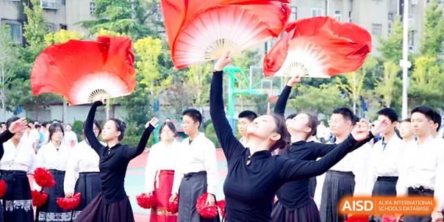 Students in black outfits perform a rhythmic fan dance with large red and white fans during an outdoor event at Zhengzhou No.19 High School.