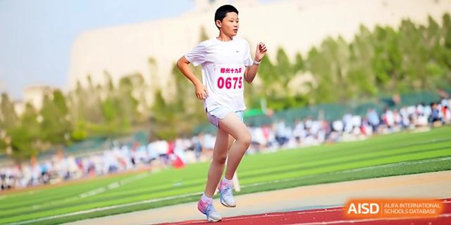 A young student athlete in a white shirt and race bib runs a track event on the green field at Zhengzhou No.19 High School.