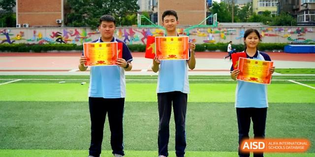 Two male students stand on the athletic field holding up their yellow award certificates in celebration at Zhengzhou No.19 High School.