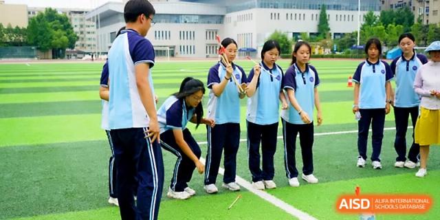 A group of female students in blue uniforms participate in a team activity or game on the school's grass field at Zhengzhou No.19 High School.