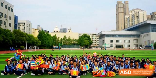 A large group of students in blue uniforms sit together on the field holding festive props and dragon decorations at Zhengzhou No.19 High School.