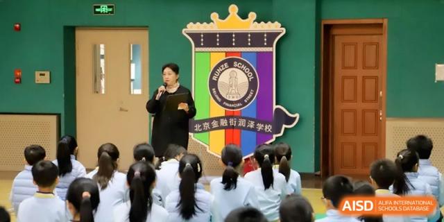 A teacher stands at a podium addressing an assembly of seated students in front of the large colorful school crest at Beijing Financial Street Runze School.