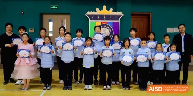 A group of elementary students stand proudly in a row holding certificates in front of a colorful school banner at Beijing Financial Street Runze School.