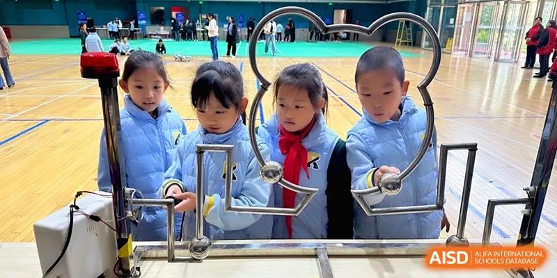 Four young students in matching blue jackets lean over a metal railing to observe a science or mechanics exhibit at Beijing Financial Street Runze School.