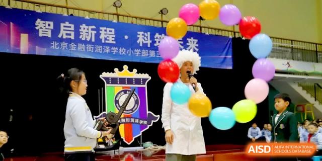 A presenter in a lab coat stands on stage decorated with colorful balloons at Beijing Financial Street Runze School.