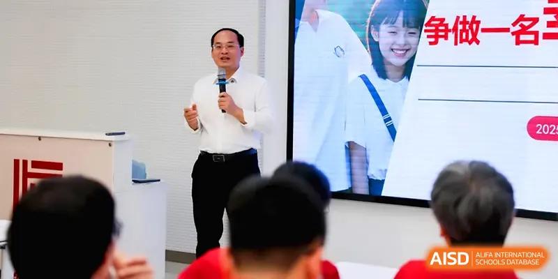 A male presenter speaks into a microphone while standing before a large digital display during a school event at Beijing Haidian Foreign Language Shi Yan School Hainan Campus.