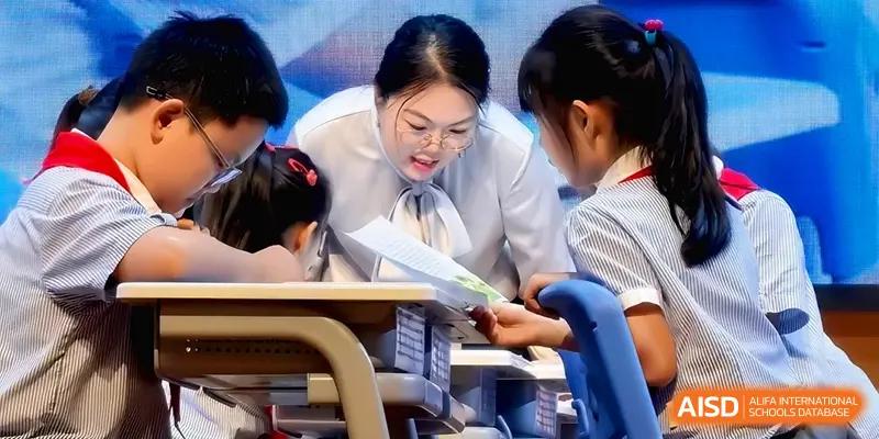 A teacher in glasses leans in to provide personalized guidance to a young student during a classroom activity at Beijing Haidian Foreign Language Shi Yan School Hainan Campus.