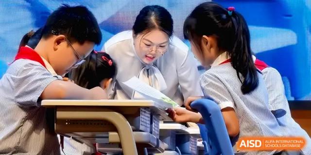 A teacher in glasses leans in to provide personalized guidance to a young student during a classroom activity at Beijing Haidian Foreign Language Shi Yan School Hainan Campus.