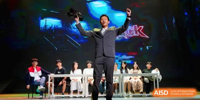 A man in a suit celebrates on stage during a performance as students watch from behind at Beijing Haidian Foreign Language Shi Yan School Hainan Campus.