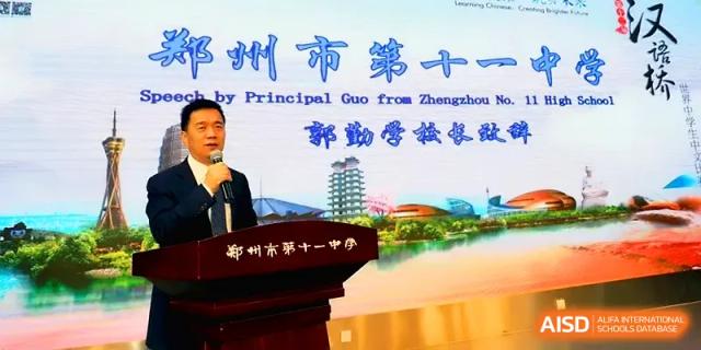 Principal Guo stands at a wooden podium giving an official speech in front of a large decorative backdrop at Zhengzhou No.11 High School.