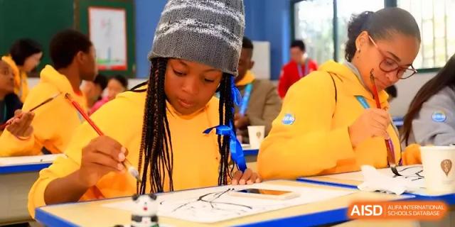 A student wearing a grey beanie and yellow shirt practices traditional Chinese calligraphy with a brush at Zhengzhou No.11 High School.