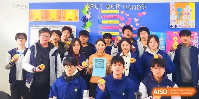 A group of smiling students and a teacher hold up handmade crafts in front of a colorful "Put Our Hands" display at Zhengzhou No.11 High School.