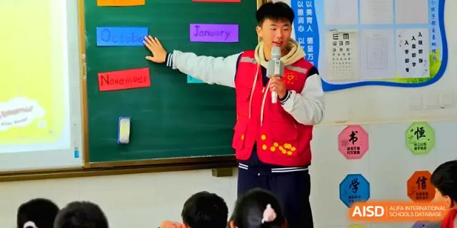 A student in a red vest stands at the front of the classroom by a chalkboard, giving a presentation at Zhengzhou No.11 High School.