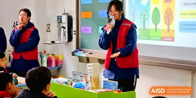 A student leader in a red and blue vest speaks into a microphone while standing at the front of a classroom at Zhengzhou No.11 High School.