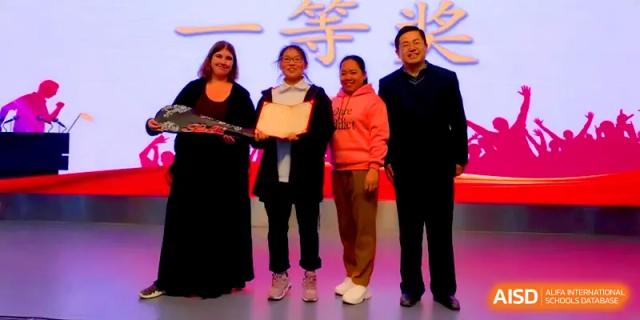 A student stands with teachers and a guest on stage while holding a certificate of achievement at Zhengzhou No.11 High School.